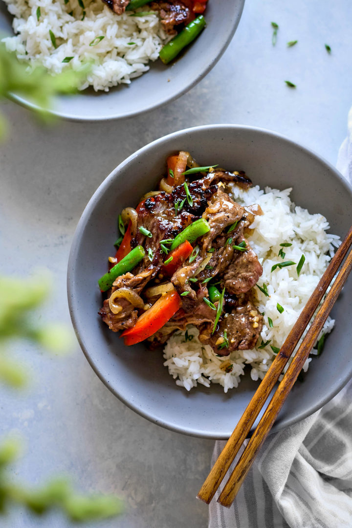 Overhead view of a serving of black pepper beef stir fry placed over white rice, plated on a blue-gray speckled plate. The beef is coated in glossy black pepper sauce with visible green beans, red bell peppers, and onions.