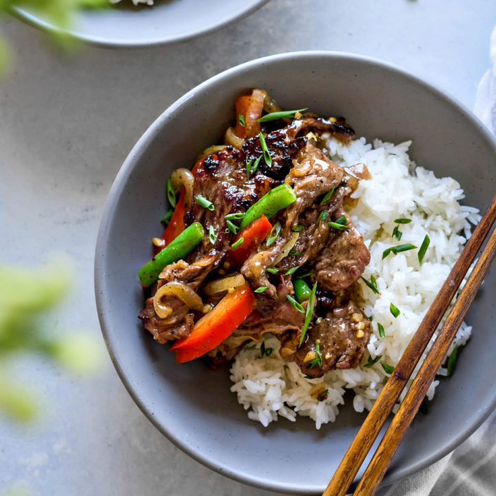 Overhead close-up of a bowl of Chinese black pepper beef served over jasmine rice in a matte grey ceramic bowl. The stir fry features seared beef strips coated in glossy black pepper sauce, tossed with red bell pepper strips, green beans, and caramelized onions. Chopped scallions are sprinkled on top. A pair of wooden chopsticks rests on the right edge of the bowl, which sits on a soft grey-and-white striped napkin. Another partially visible bowl appears in the top left corner, and a blurred leafy green element is in the foreground.