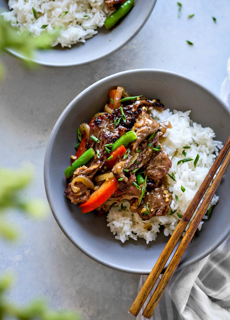 Overhead close-up of a bowl of Chinese black pepper beef served over jasmine rice in a matte grey ceramic bowl. The stir fry features seared beef strips coated in glossy black pepper sauce, tossed with red bell pepper strips, green beans, and caramelized onions. Chopped scallions are sprinkled on top. A pair of wooden chopsticks rests on the right edge of the bowl, which sits on a soft grey-and-white striped napkin. Another partially visible bowl appears in the top left corner, and a blurred leafy green element is in the foreground.