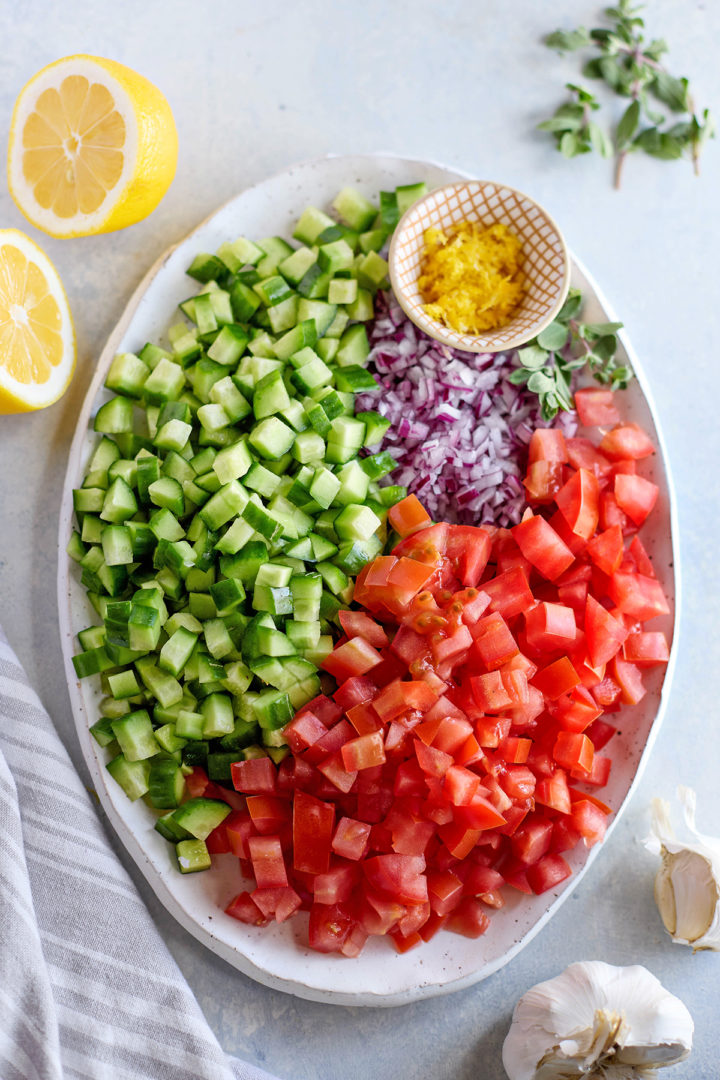 An overhead view of a white oval platter holding neatly chopped fresh vegetables for Greek salad. From left to right: diced cucumber, finely chopped red onion, and diced tomato. A small bowl of bright yellow lemon zest sits in the center, surrounded by a few sprigs of fresh oregano. To the side are halved lemons and two garlic cloves.
