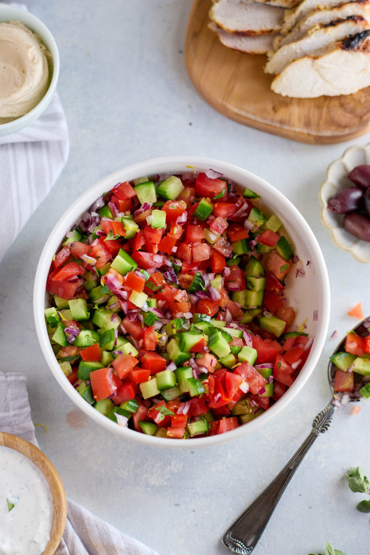 A white bowl is filled with a freshly made cucumber tomato salad, featuring small diced pieces of red tomato, green cucumber, and purple onion. The salad is mixed and slightly glossy, suggesting a light dressing. Surrounding the bowl are key components of the meal: a small bowl of hummus on the left, sliced grilled chicken on a wooden board in the top right, and a dish of Kalamata olives nearby. A silver spoon rests beside the salad, and a dollop of tzatziki is visible in a wooden bowl at the bottom edge of the frame.