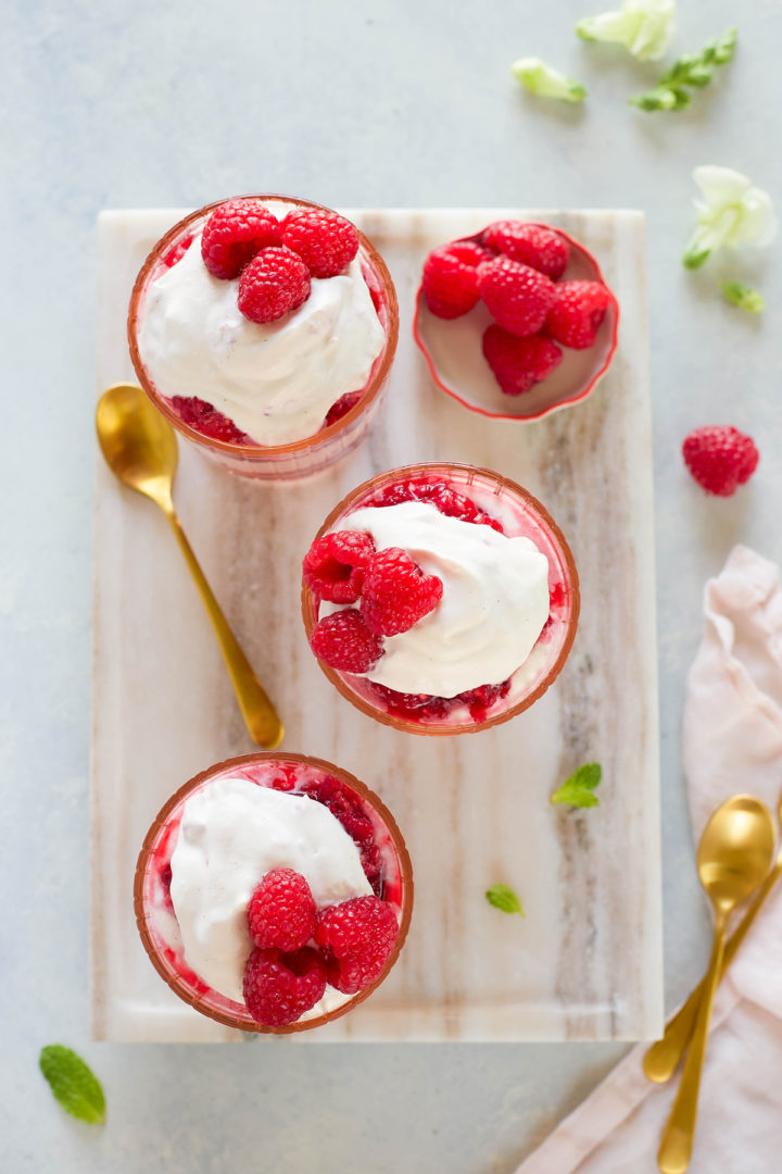 Three servings of raspberry fool dessert layered with crushed raspberries and whipped cream in pink glasses, each topped with fresh raspberries. A gold spoon and scattered mint leaves complete the scene.