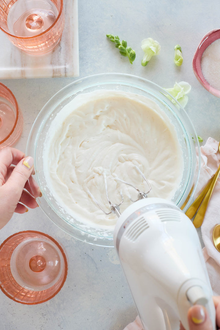 Overhead view of a glass bowl filled with whipped cream being mixed with a hand mixer, part of the preparation for an easy raspberry fool recipe. Surrounding elements include pink dessert glasses and gold spoons.