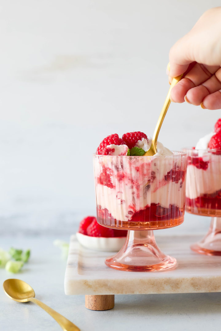 Hand holding a gold spoon digging into a glass of raspberry fool dessert, showing layers of raspberries and whipped cream with a soft swirl texture.