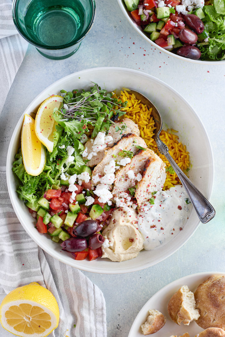 Close-up of colorful Greek chicken bowls featuring seasoned grilled chicken over turmeric rice, topped with tzatziki, fresh vegetables, and feta cheese.