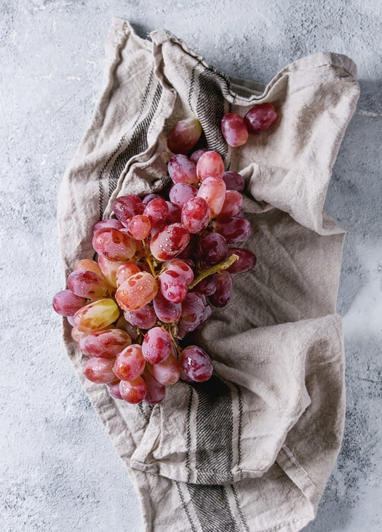 A freshly rinsed bunch of red grapes rests on a soft beige and gray-striped kitchen towel, laid out on a textured light gray surface. The grapes are plump with visible water droplets, indicating they’ve just been washed and are in the process of being dried before freezing.