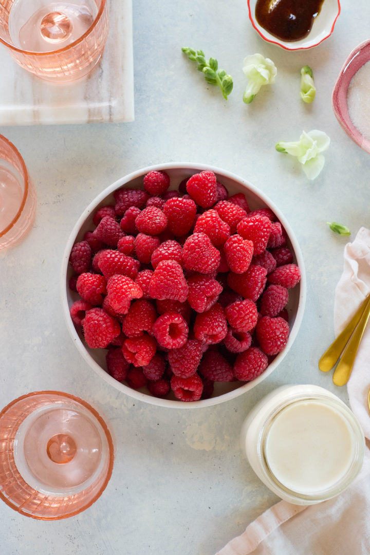 Large bowl of fresh raspberries on a light surface with other raspberry fool ingredients nearby, including cream, sugar, and glass serving dishes.