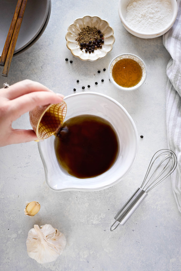 Close-up photo of a woman combining ingredients together in a small white bowl to make the black pepper sauce. She holds is pouring the coconut aminos into a bowl with other ingredients and has other small ingredient bowls nearby.