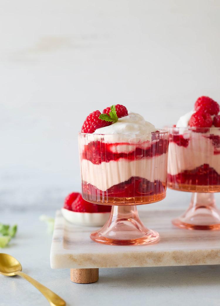 Layered raspberry fool dessert served in vintage-style pink glass cups, with swirls of whipped cream and raspberry compote, garnished with fresh berries and mint on a marble serving board.