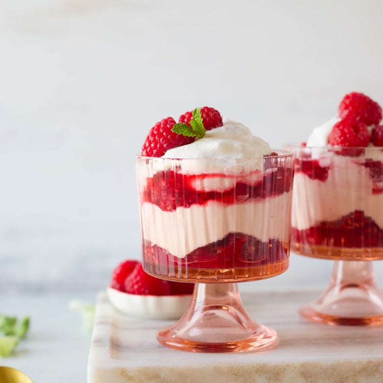 Layered raspberry fool dessert served in vintage-style pink glass cups, with swirls of whipped cream and raspberry compote, garnished with fresh berries and mint on a marble serving board.