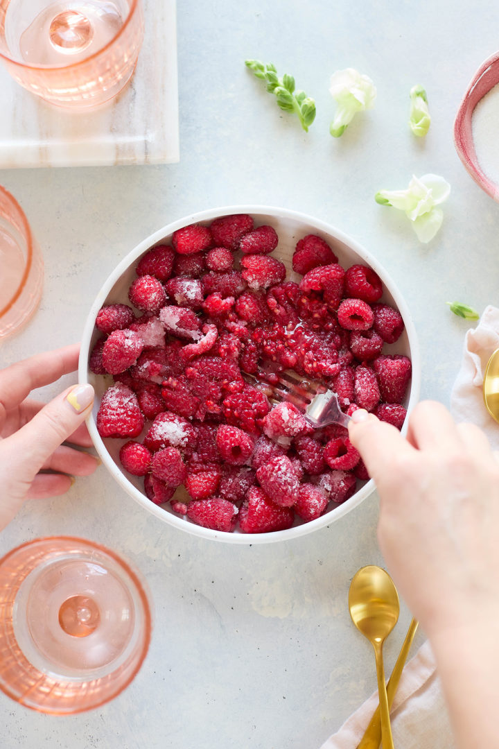 Close-up of raspberries being mashed in a white bowl with sugar using a fork, part of the process for preparing a raspberry fool dessert.
