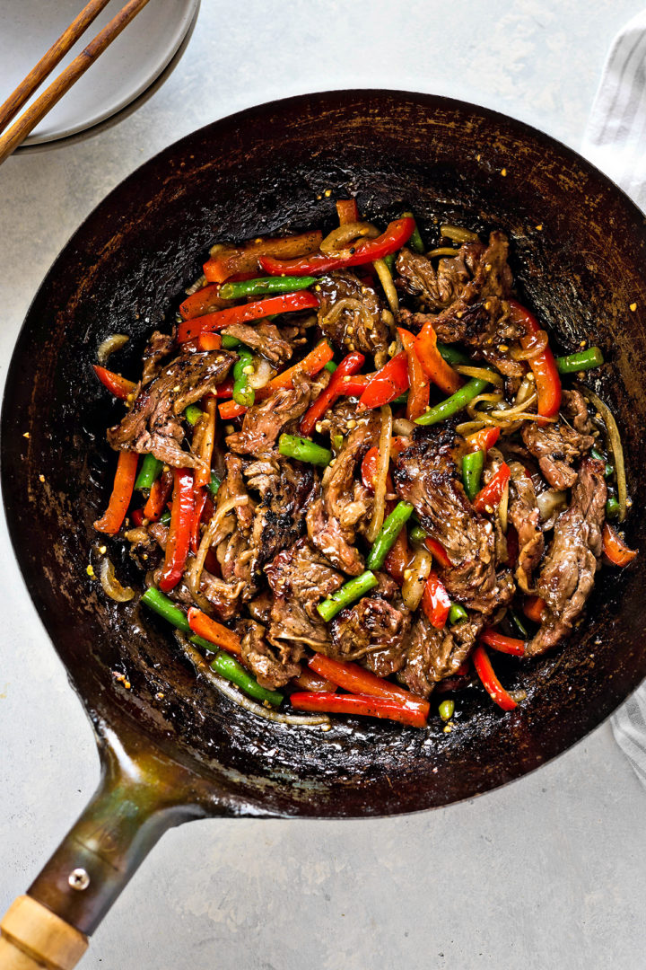 Action shot of a wok containing seared beef with black pepper sauce, red bell peppers, green beans, and onions. The ingredients are just finished being stir fried.
