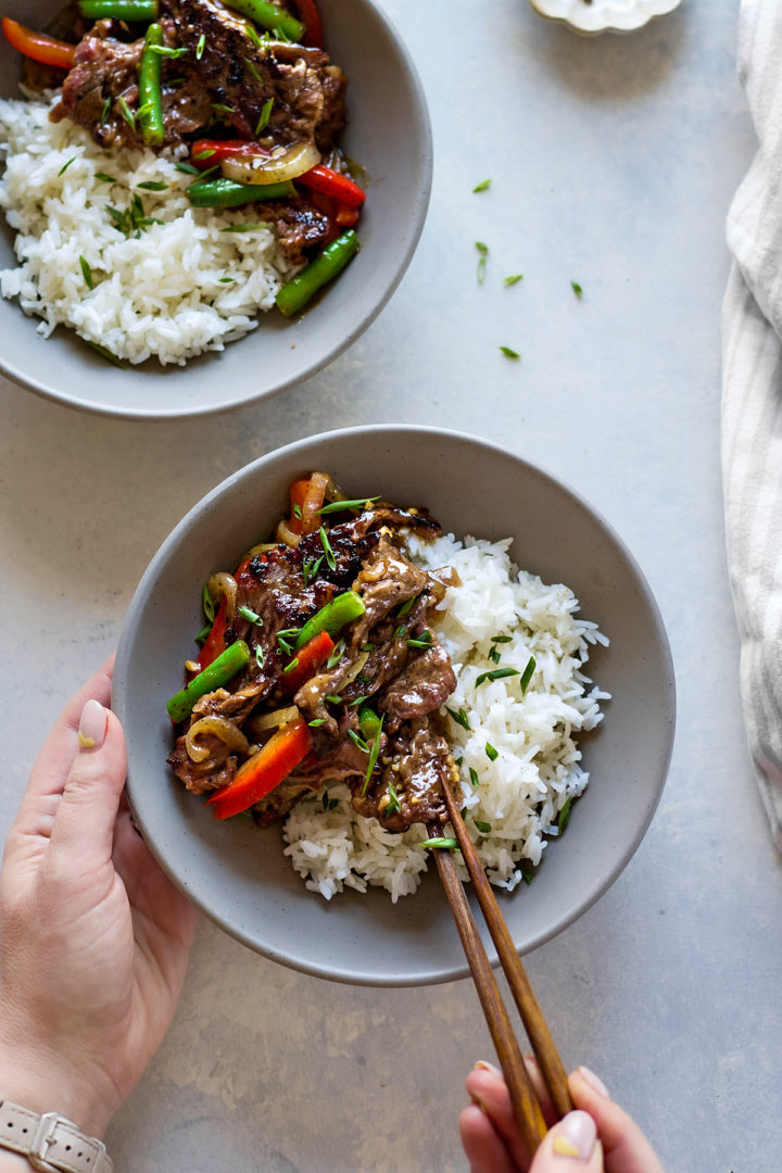 Overhead view of a woman eating a bowl of Chinese black pepper beef served on a grey ceramic plate. The beef and vegetables are glossy and saucy, with black pepper visibly flecked across the dish. Fluffy white jasmine rice sits beside the stir fry on the plate.