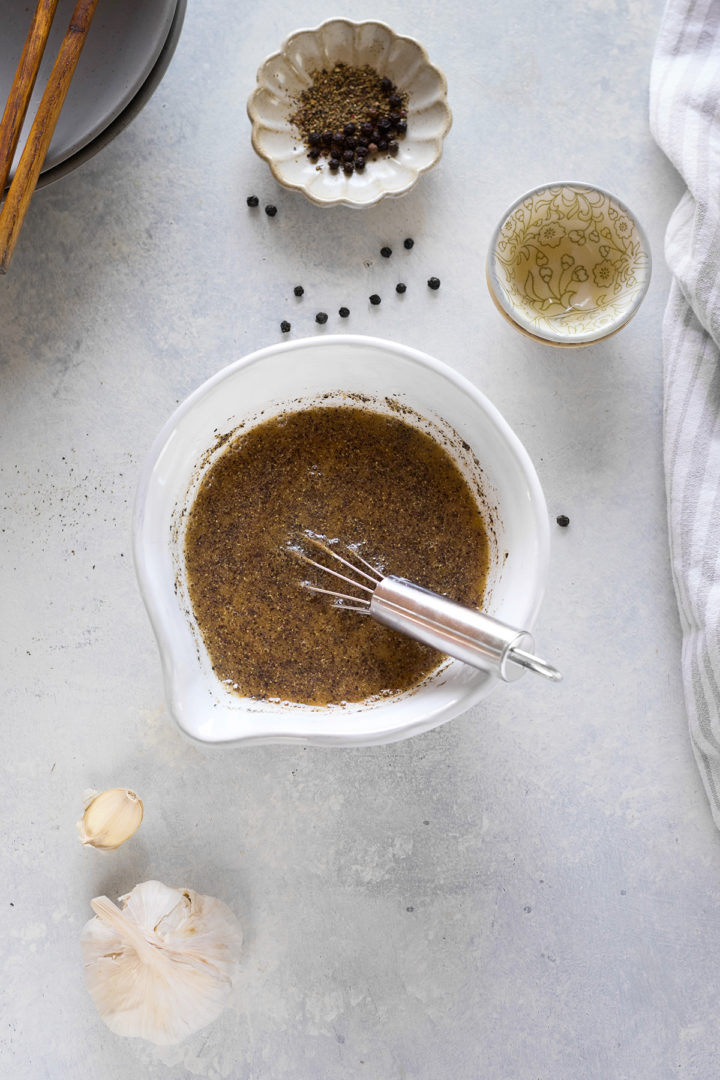 Overhead view of a small white mixing bowl filled with prepared black pepper sauce, with a metal whisk resting inside. The sauce is dark and speckled with visible black pepper. Surrounding the bowl on the light textured surface are a head of garlic, a garlic clove, a small fluted dish of coarsely ground black pepper and peppercorns, and a small glass bowl of dry sherry. A folded grey-and-white striped cloth is partially visible on the right edge.