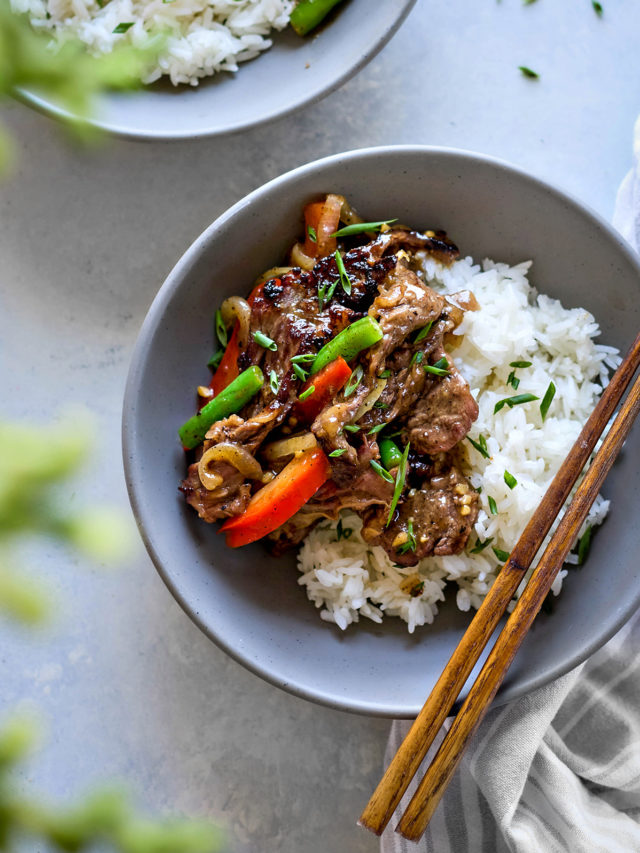 Overhead close-up of a bowl of Chinese black pepper beef served over jasmine rice in a matte grey ceramic bowl. The stir fry features seared beef strips coated in glossy black pepper sauce, tossed with red bell pepper strips, green beans, and caramelized onions. Chopped scallions are sprinkled on top. A pair of wooden chopsticks rests on the right edge of the bowl, which sits on a soft grey-and-white striped napkin. Another partially visible bowl appears in the top left corner, and a blurred leafy green element is in the foreground.