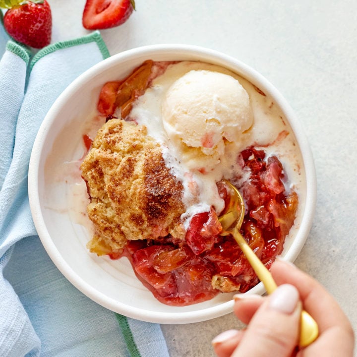 A close-up of a hand holding a gold spoon, scooping into a warm serving of strawberry rhubarb cobbler with biscuit topping and vanilla ice cream, in a white bowl on a pale background.