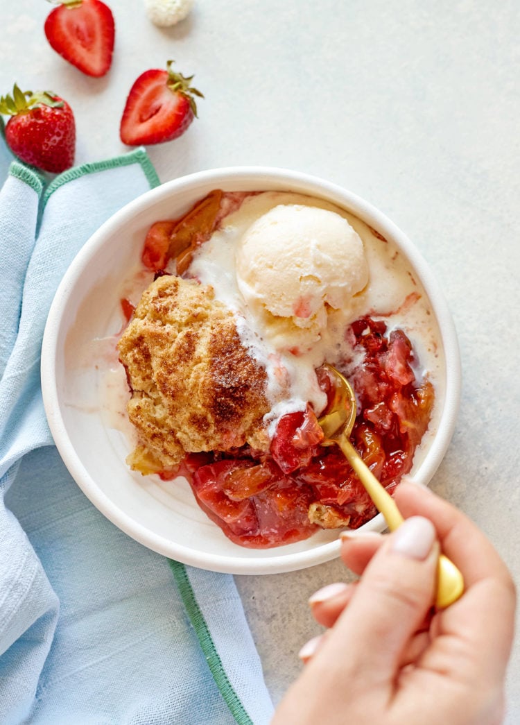 A close-up of a hand holding a gold spoon, scooping into a warm serving of strawberry rhubarb cobbler with biscuit topping and vanilla ice cream, in a white bowl on a pale background.