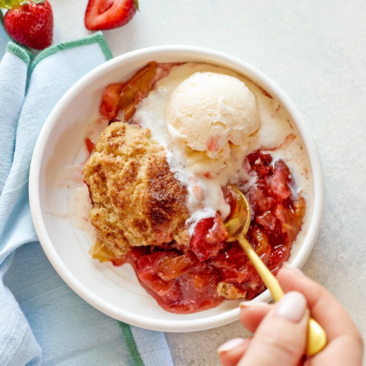 A close-up of a hand holding a gold spoon, scooping into a warm serving of strawberry rhubarb cobbler with biscuit topping and vanilla ice cream, in a white bowl on a pale background.