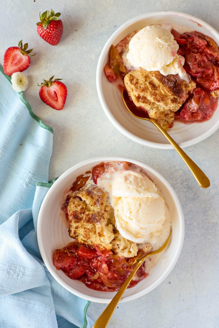 Overhead view of two white bowls filled with warm strawberry rhubarb cobbler topped with golden-brown biscuit topping and scoops of vanilla ice cream. The bowls sit on a light surface with a gold spoon in each, surrounded by fresh strawberries and a folded blue napkin.
