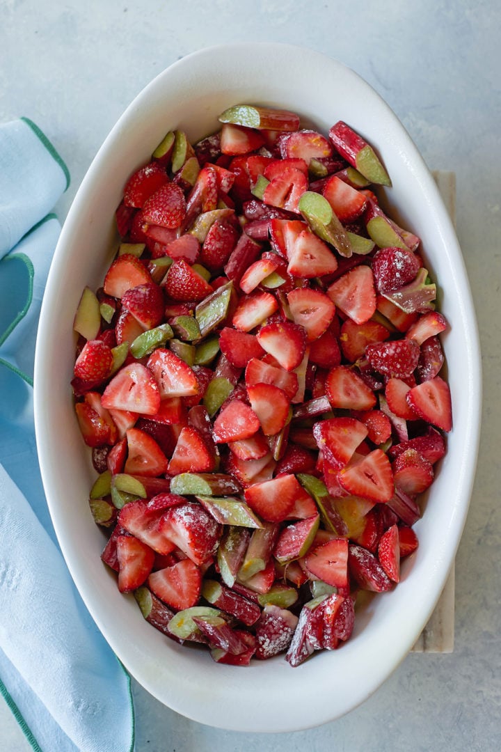 An oval white baking dish filled with raw, sliced strawberries and rhubarb, lightly coated in sugar. The fruit is vibrant and fresh, resting on a light blue surface with a soft napkin beside the dish.