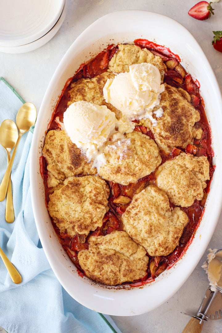 Overhead view of a fully baked strawberry rhubarb cobbler in a white oval dish. The cobbler has golden biscuit-style topping and is served with several scoops of melting vanilla ice cream on top. A few strawberries and gold spoons are visible nearby.