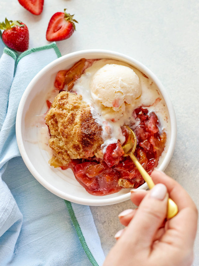 A close-up of a hand holding a gold spoon, scooping into a warm serving of strawberry rhubarb cobbler with biscuit topping and vanilla ice cream, in a white bowl on a pale background.