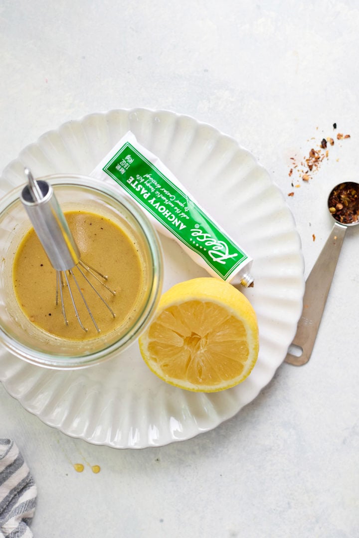 A jar of Caesar chicken marinade being whisked on a white scalloped plate. A halved lemon, red pepper flakes, and a tube of anchovy paste are nearby on a pale surface.
