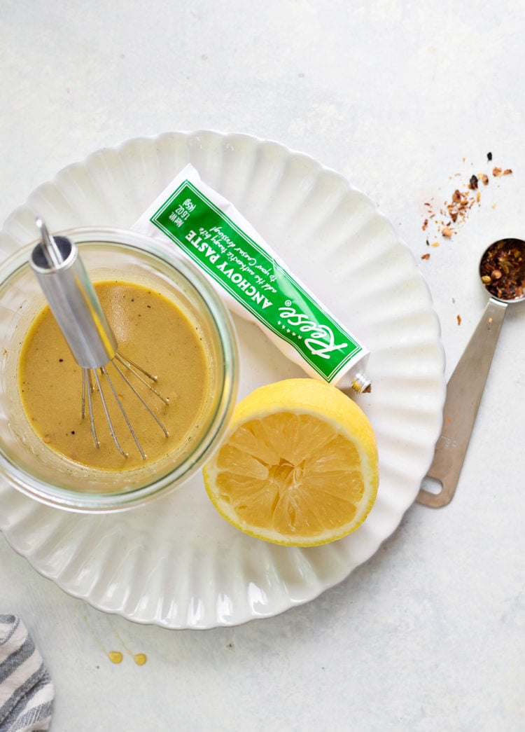 A jar of Caesar chicken marinade being whisked on a white scalloped plate. A halved lemon, red pepper flakes, and a tube of anchovy paste are nearby on a pale surface.