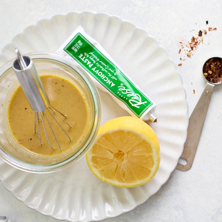 A jar of Caesar chicken marinade being whisked on a white scalloped plate. A halved lemon, red pepper flakes, and a tube of anchovy paste are nearby on a pale surface.