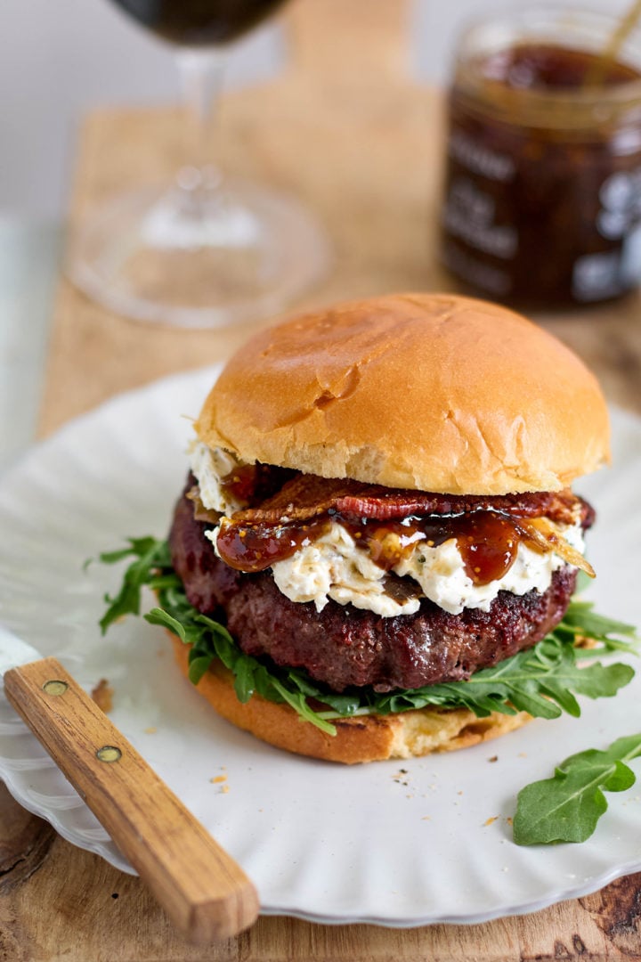 Close-up of a goat cheese burger on a white plate, layered with arugula, a juicy grilled patty, creamy goat cheese, fig jam, and crispy bacon on a toasted bun.
