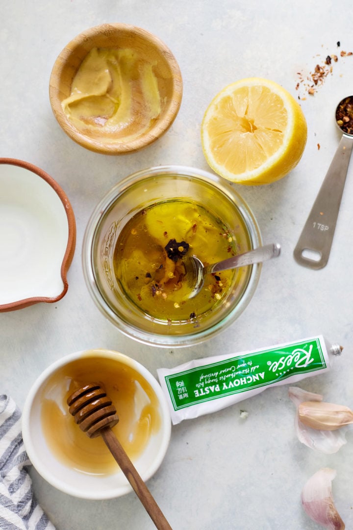 A close-up of Caesar chicken marinade ingredients being mixed in a glass jar. Surrounding the bowl are a halved lemon, a small bowl of mustard, garlic cloves, honey, anchovy paste, and red pepper flakes.
