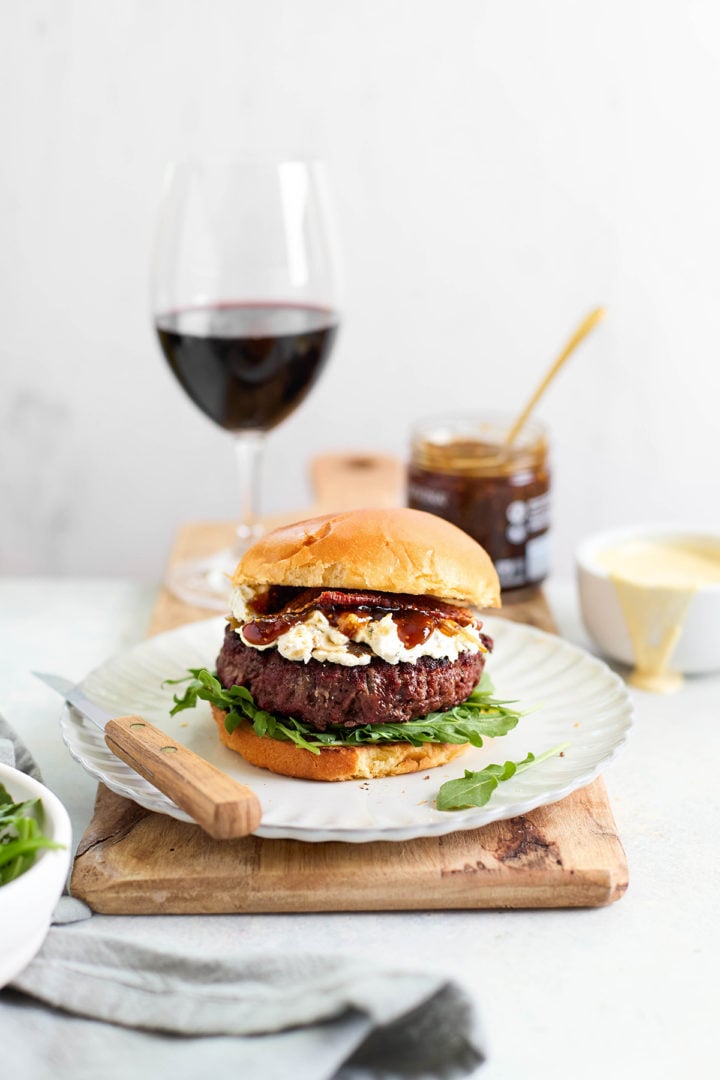 A finished goat cheese burger placed on a rustic wood board, layered with arugula, bacon, fig jam, and herbed goat cheese spread between toasted buns.