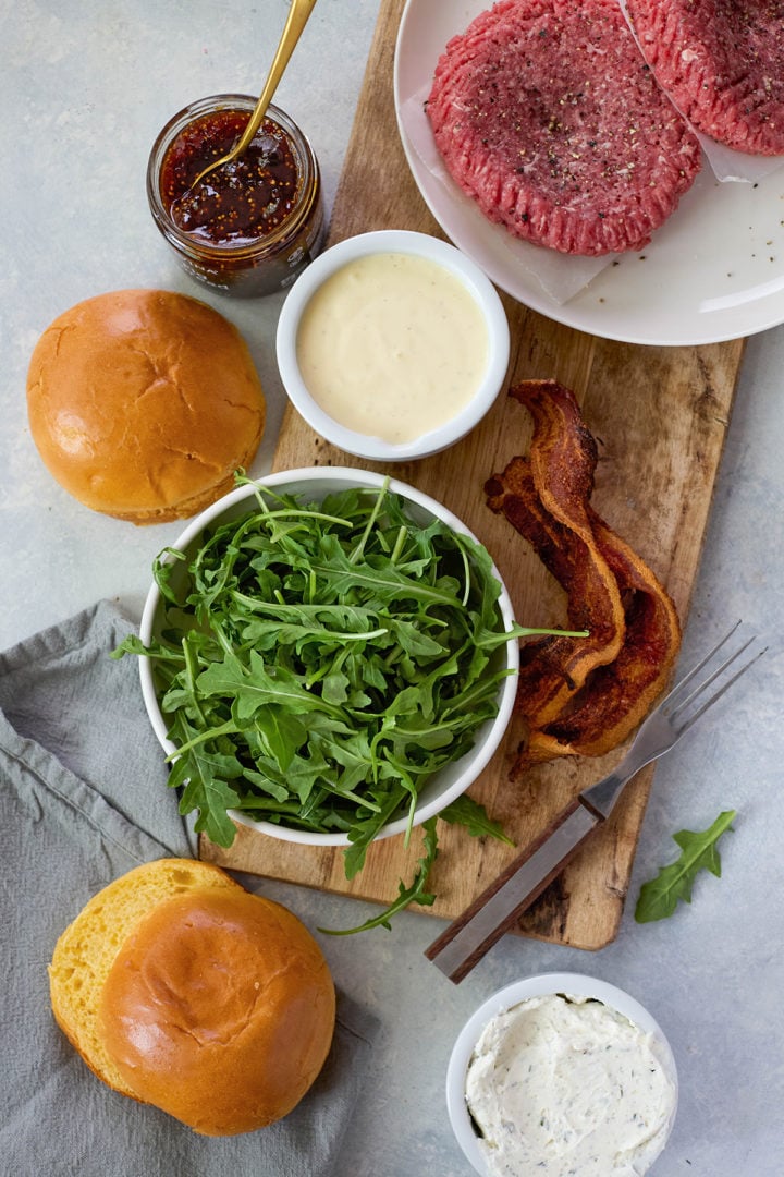 Overhead shot of ingredients for a goat cheese burger, including arugula, bacon strips, burger patties, fig jam, garlic herb goat cheese, sauce, and brioche buns on a cutting board.
