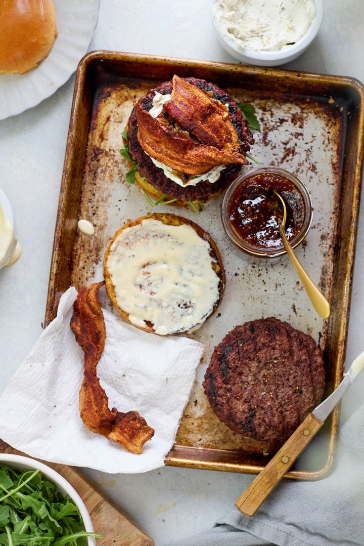 Head-on view of a goat cheese burger with arugula, creamy goat cheese, crispy bacon, and glossy fig jam, stacked on a toasted bun and served on a scalloped white plate.
