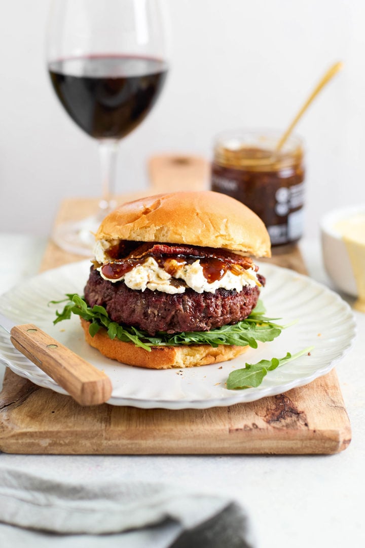 Burger on a white plate with arugula, goat cheese, bacon, and fig jam, with wine and a jar of jam in the background — styled for a gourmet meal setting.
