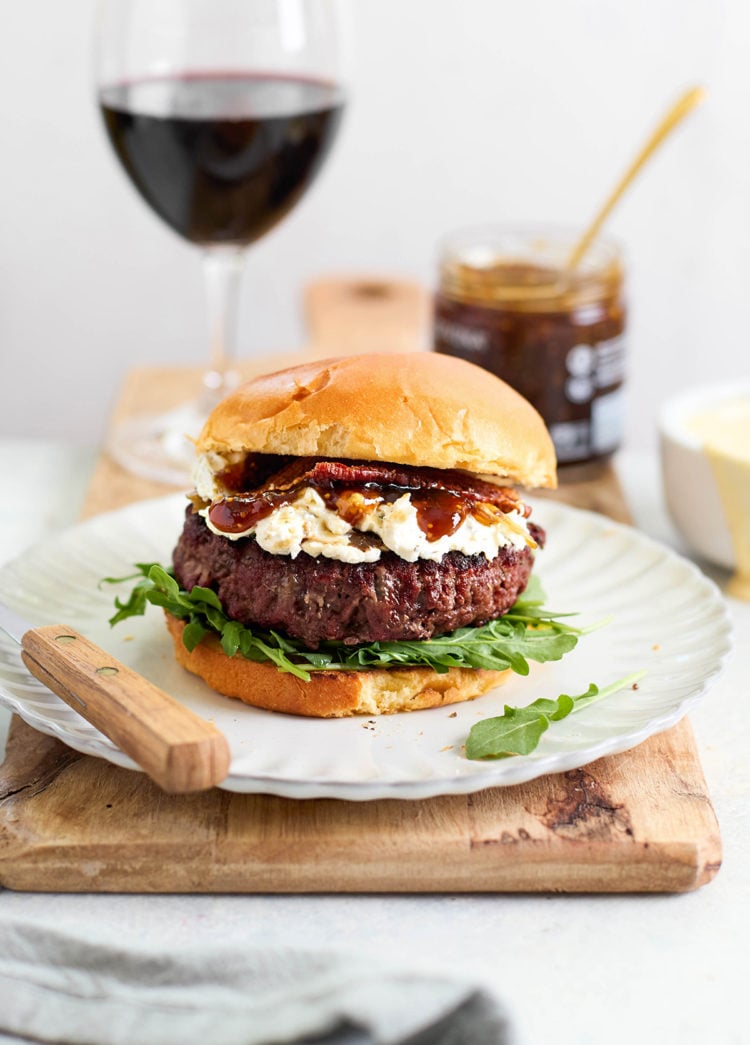 Burger on a white plate with arugula, goat cheese, bacon, and fig jam, with wine and a jar of jam in the background — styled for a gourmet meal setting.