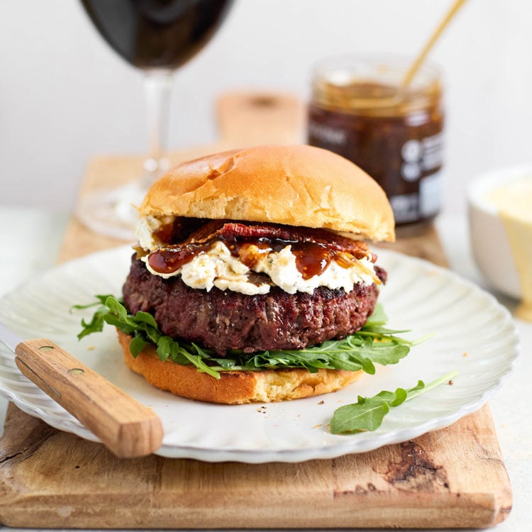 Burger on a white plate with arugula, goat cheese, bacon, and fig jam, with wine and a jar of jam in the background — styled for a gourmet meal setting.