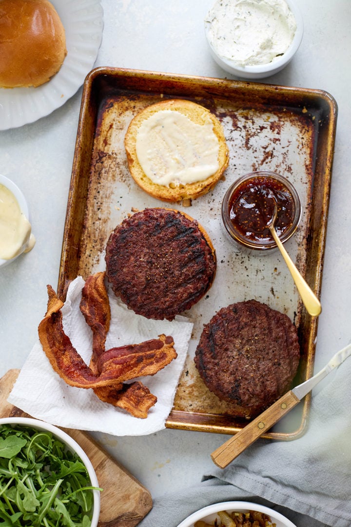 Overhead view of burger assembly on a sheet pan with grilled patties, fig jam, bacon, mayonnaise, arugula, and herbed goat cheese being spooned onto a burger.

