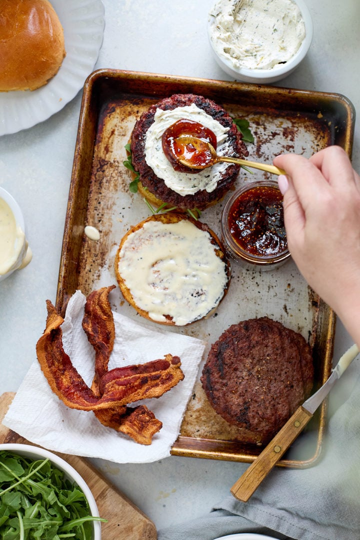 Flat lay of two cooked burger patties, fig jam, toasted buns, bacon, and arugula on a sheet pan — ingredients laid out for assembling goat cheese burgers.
