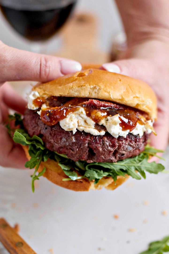 A hand holding a goat cheese burger with layers of arugula, grilled beef patty, whipped goat cheese, fig jam, and bacon, mid-serve over a plate.

