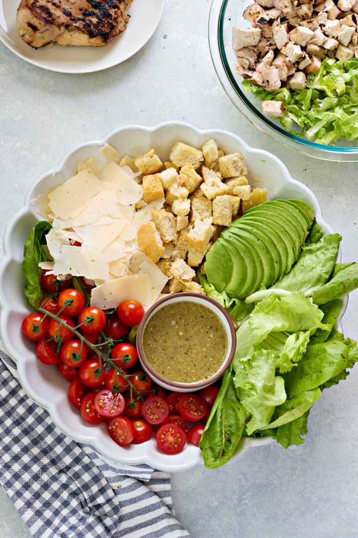 A white scalloped dish filled with Caesar salad ingredients: cherry tomatoes on the vine, shaved parmesan, croutons, romaine lettuce, sliced avocado, and Caesar chicken marinade in a small bowl. Grilled and chopped chicken are visible in nearby bowls.