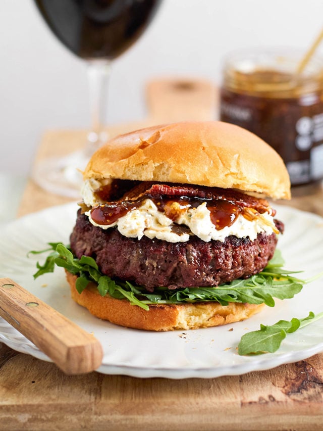 Burger on a white plate with arugula, goat cheese, bacon, and fig jam, with wine and a jar of jam in the background — styled for a gourmet meal setting.