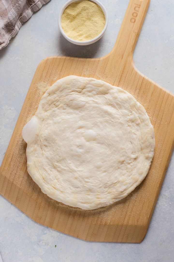  Unbaked pizza dough spread with tomato sauce and topped with torn mozzarella. Bowls of fresh arugula, prosciutto slices, and mozzarella are arranged beside the pizza on a wooden peel.