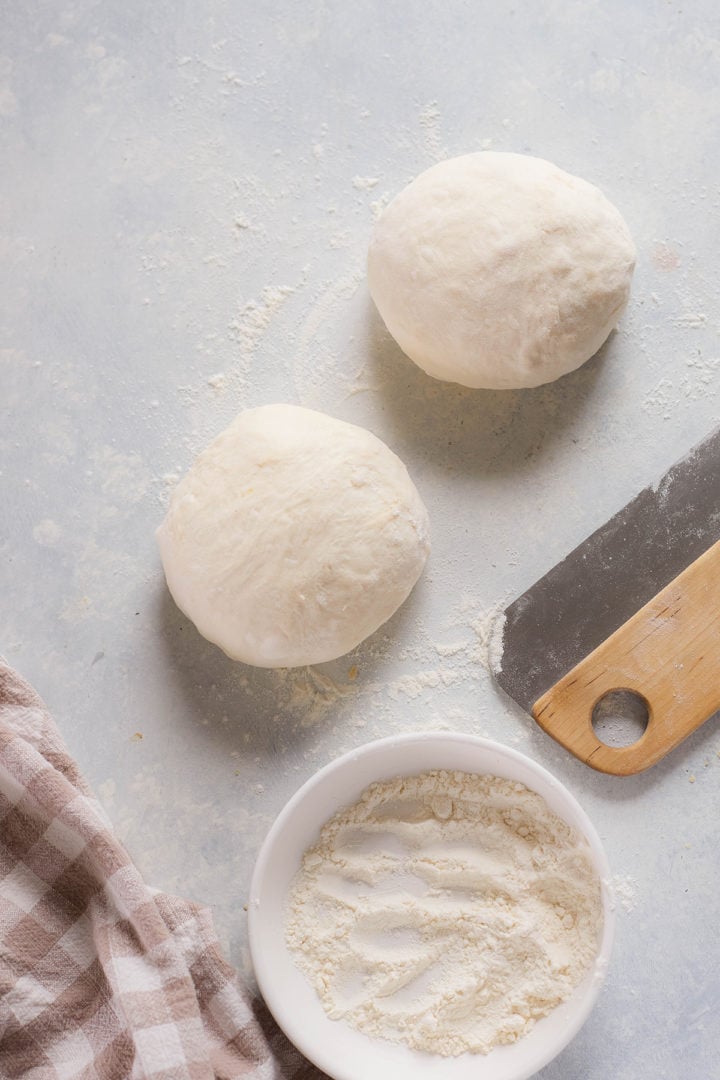 Two balls of homemade pizza dough on a floured surface with a bowl of flour and a bench scraper nearby.