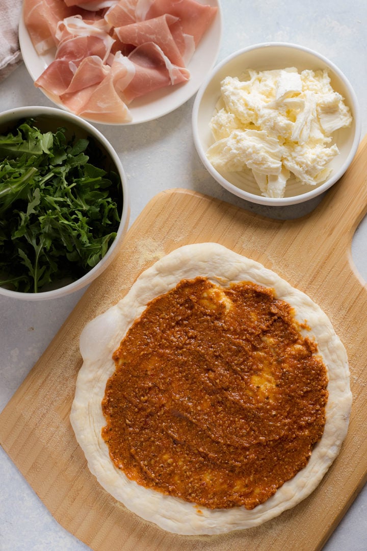 Unbaked pizza dough spread with tomato sauce and topped with torn mozzarella. Bowls of fresh arugula, prosciutto slices, and mozzarella are arranged beside the pizza on a wooden peel.