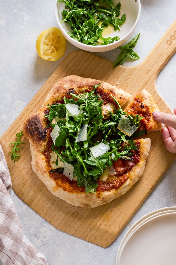 A baked pizza on a wooden peel topped with fresh arugula, shaved Parmesan, and tomato sauce. A hand is pulling away one slice, showing the crisp crust. A bowl of dressed arugula salad and half a lemon sit nearby.