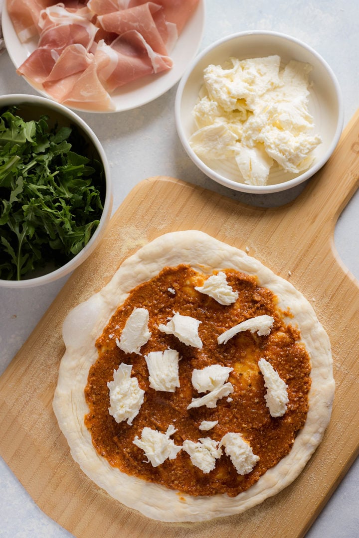 Unbaked pizza dough spread with tomato sauce and topped with torn mozzarella. Bowls of fresh arugula, prosciutto slices, and mozzarella are arranged beside the pizza on a wooden peel.