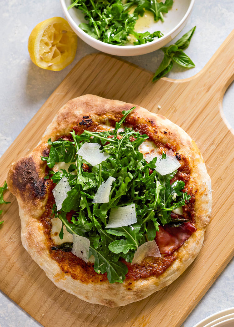 Freshly baked pizza on a wooden board, topped with arugula salad and Parmesan shavings. A bowl of arugula salad with vinaigrette and a squeezed lemon are in the background.