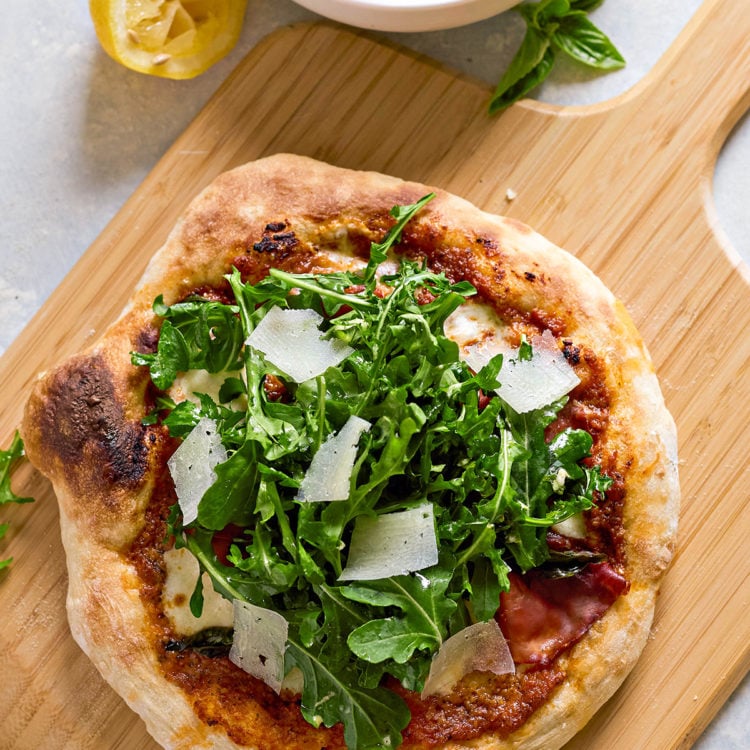 Freshly baked pizza on a wooden board, topped with arugula salad and Parmesan shavings. A bowl of arugula salad with vinaigrette and a squeezed lemon are in the background.