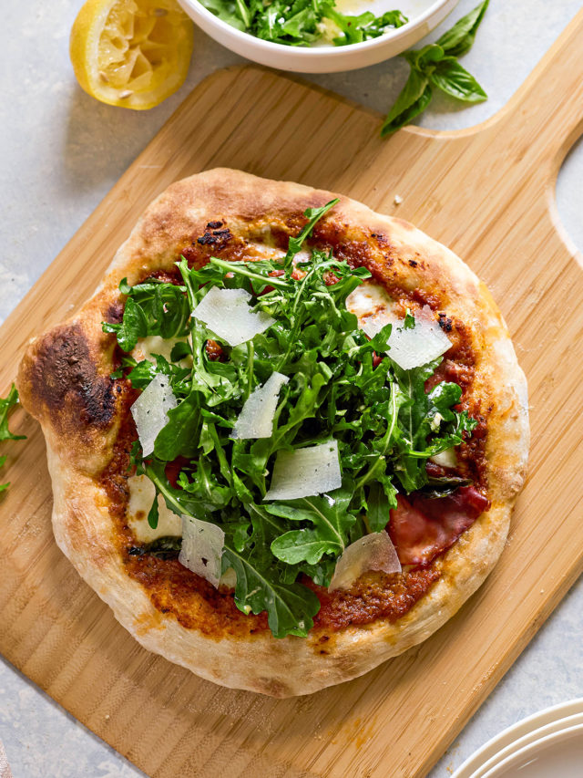 Freshly baked pizza on a wooden board, topped with arugula salad and Parmesan shavings. A bowl of arugula salad with vinaigrette and a squeezed lemon are in the background.